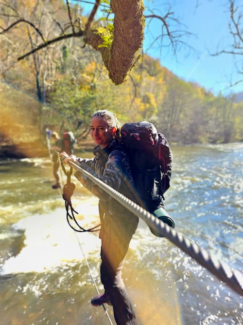 Vrouw steekt een rivier over via een touwbrug tijdens een survival- en bushcraftcursus, met een grote rugzak en outdooruitrusting in een bosrijke omgeving.