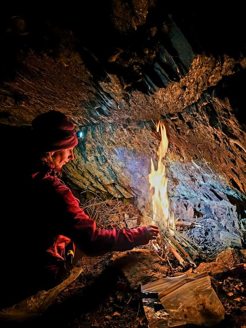 Persoon maakt ’s nachts een vuur onder een rotswand tijdens een survival- en bushcraftcursus, met natuurlijke materialen en een hoofdlamp voor zicht
