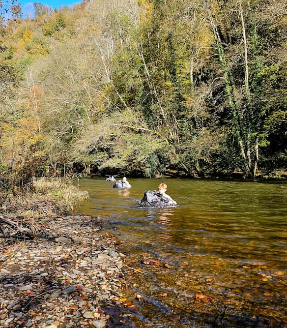 Survivaldeelnemers steken een koude rivier over met hun rugzakken boven water.