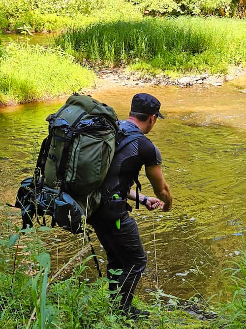 Een man met een grote rugzak vist in een ondiepe beek tijdens een survivaltocht.