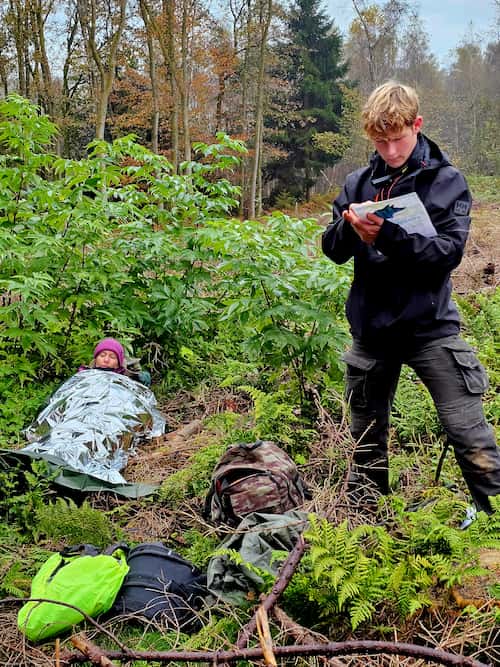 Een jongere maakt aantekeningen terwijl een ander in een nooddeken ligt tijdens een survivaloefening.
